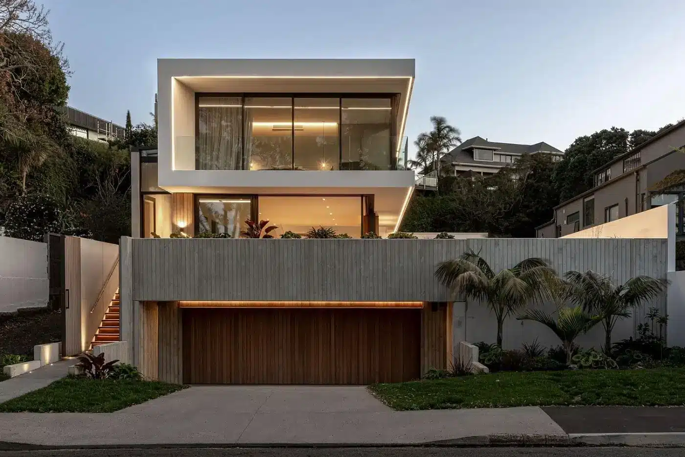 A three-story contemporary home featuring white cantilevered slabs, floor-to-ceiling glass, and a natural wood garage door set against a concrete retaining wall.