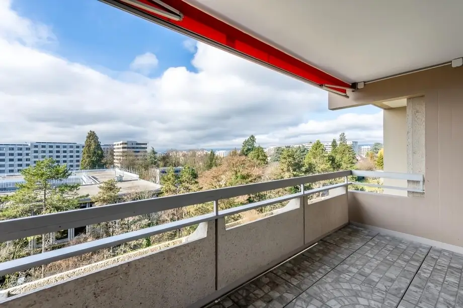 Un balcon spacieux avec un sol carrelé gris, une balustrade en métal et un auvent rouge rétractable donnant sur un parc urbain.