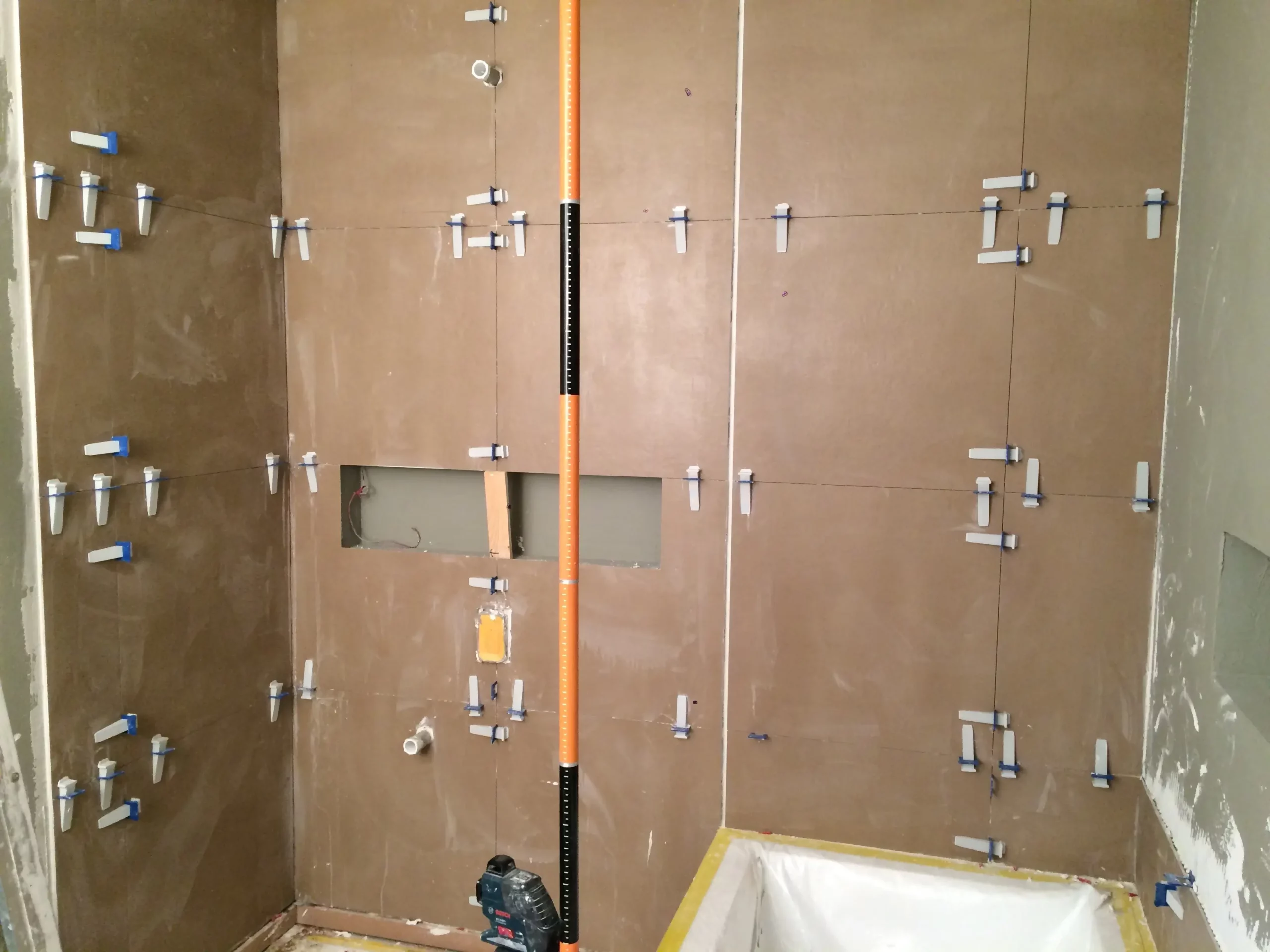 Grands carreaux muraux marron en cours d'installation dans une zone de douche à l'aide d'un système d'entretoise de nivellement et d'un niveau laser.