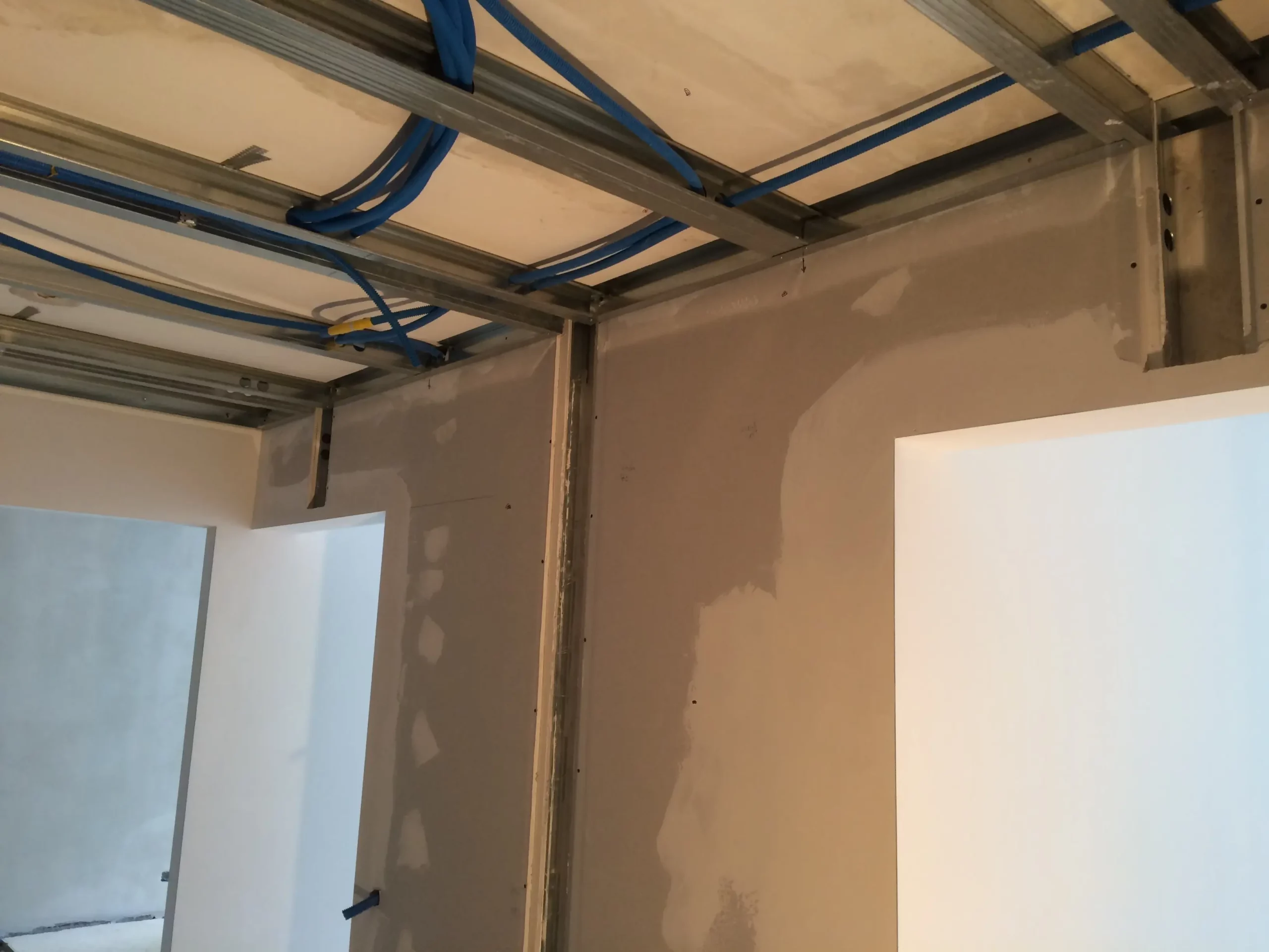 Un chantier de construction montrant des rails de plafond en métal avec des conduits électriques bleus et des cloisons en plaques de plâtre plâtrées.